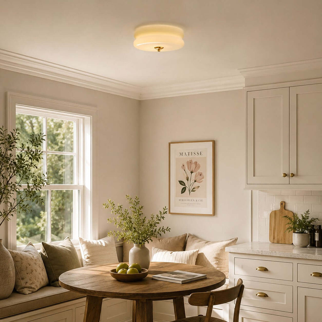 This Clérachic flush mount ceiling light glows over a breakfast nook with a window seat, a round table and a framed floral print.
