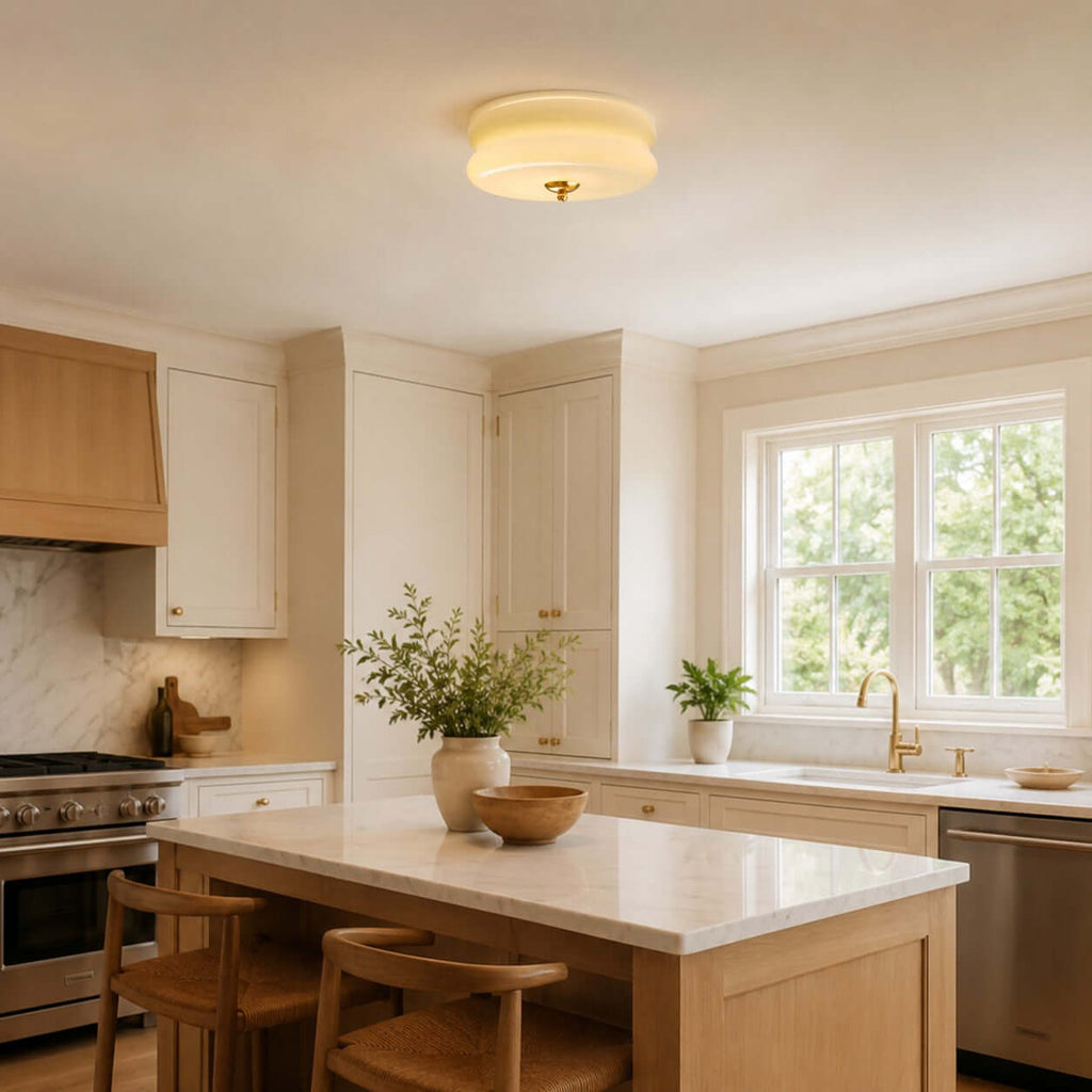 A Clérachic layered glass ceiling light sits flush above a kitchen with cream cabinets, a wooden island and woven stools.