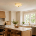 A Clérachic layered glass ceiling light sits flush above a kitchen with cream cabinets, a wooden island and woven stools.