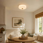 A Clérachic flush mount with a soft white glass shade lights a dining corner with a round table, a built-in bench and a landscape print.