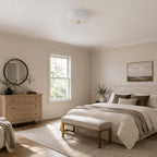 This unlit white Clérachic flush mount sits on the ceiling of a bedroom with a pale chest of drawers, a round mirror and a bench.
