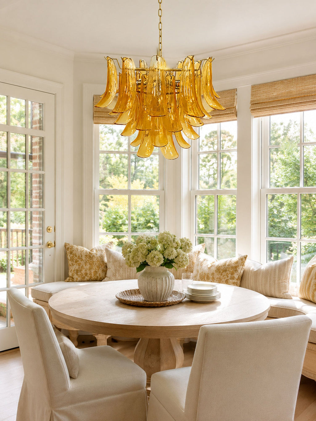 This Clérachic amber glass chandelier hangs in a bay window over a round pedestal table and cream dining chairs.