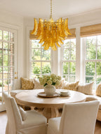 This Clérachic amber glass chandelier hangs in a bay window over a round pedestal table and cream dining chairs.