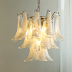 Milky white glass leaves of this Clérachic chandelier glow with a warm light, set against a grey wall and a sheer curtain.