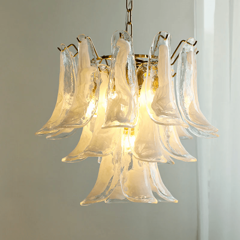 Milky white glass leaves of this Clérachic chandelier glow with a warm light, set against a grey wall and a sheer curtain.