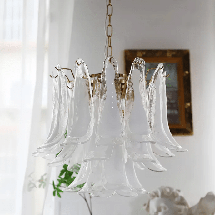 This Clérachic clear glass leaf chandelier hangs near a gilt-framed mirror, with sheer curtains and a small plaster figure below.