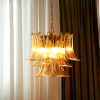 This Clérachic amber glass chandelier glows between sheer white curtains and a heavy brown drape, its brass ring just visible.