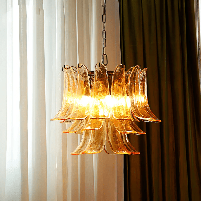 This Clérachic amber glass chandelier glows between sheer white curtains and a heavy brown drape, its brass ring just visible.