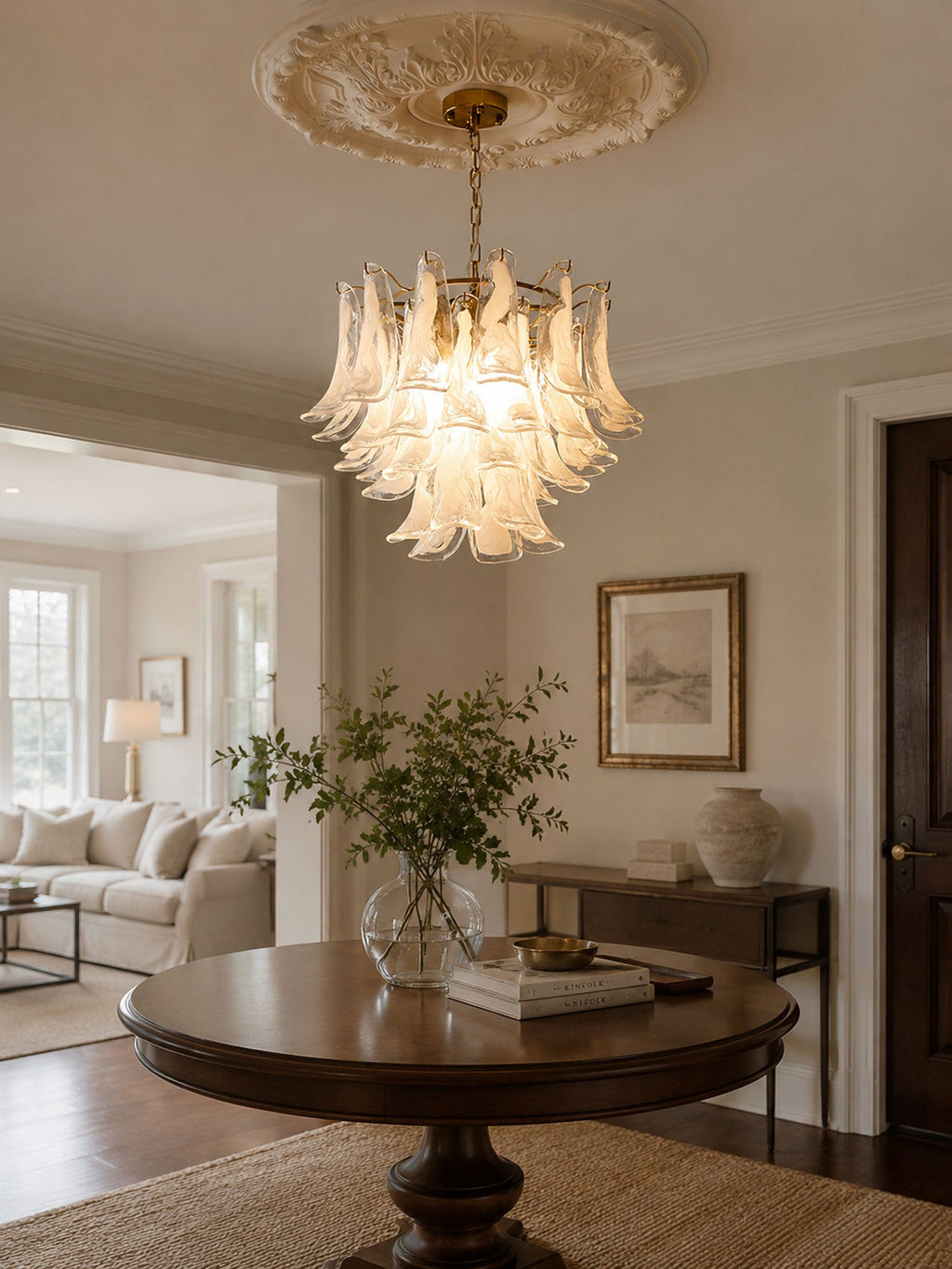 Frosted white glass chandelier from Clérachic hanging in a hall below a ceiling rose, above a round table with a plant in a vase.
