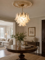 Frosted white glass chandelier from Clérachic hanging in a hall below a ceiling rose, above a round table with a plant in a vase.