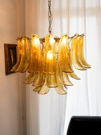 Amber glass leaf chandelier from Clérachic, lit and seen at a slight angle beside a framed portrait and sheer curtains.