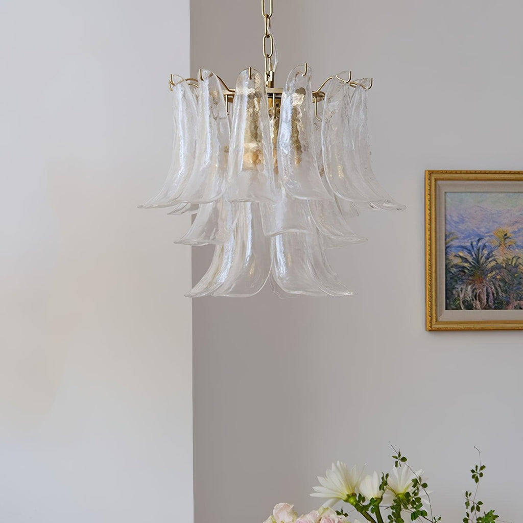 Slim clear glass leaf chandelier from Clérachic against a white wall, with a framed landscape and white flowers at the edge.
