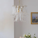 Slim clear glass leaf chandelier from Clérachic against a white wall, with a framed landscape and white flowers at the edge.