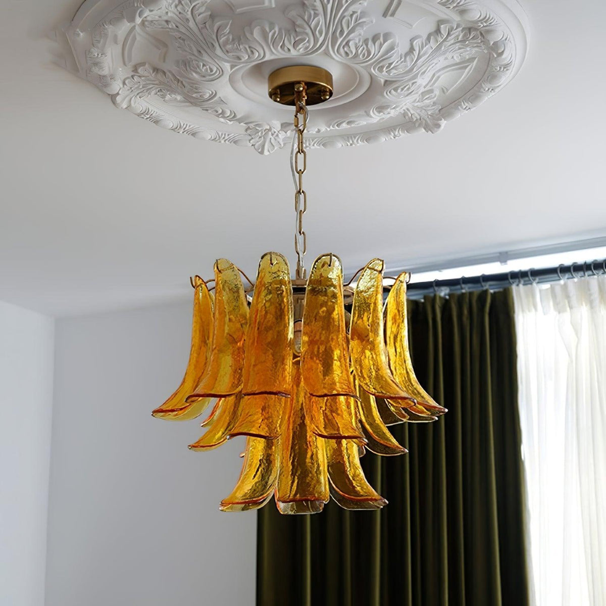 Unlit amber glass leaf chandelier from Clérachic beneath a plaster ceiling rose, in front of green curtains and sheer drapes.
