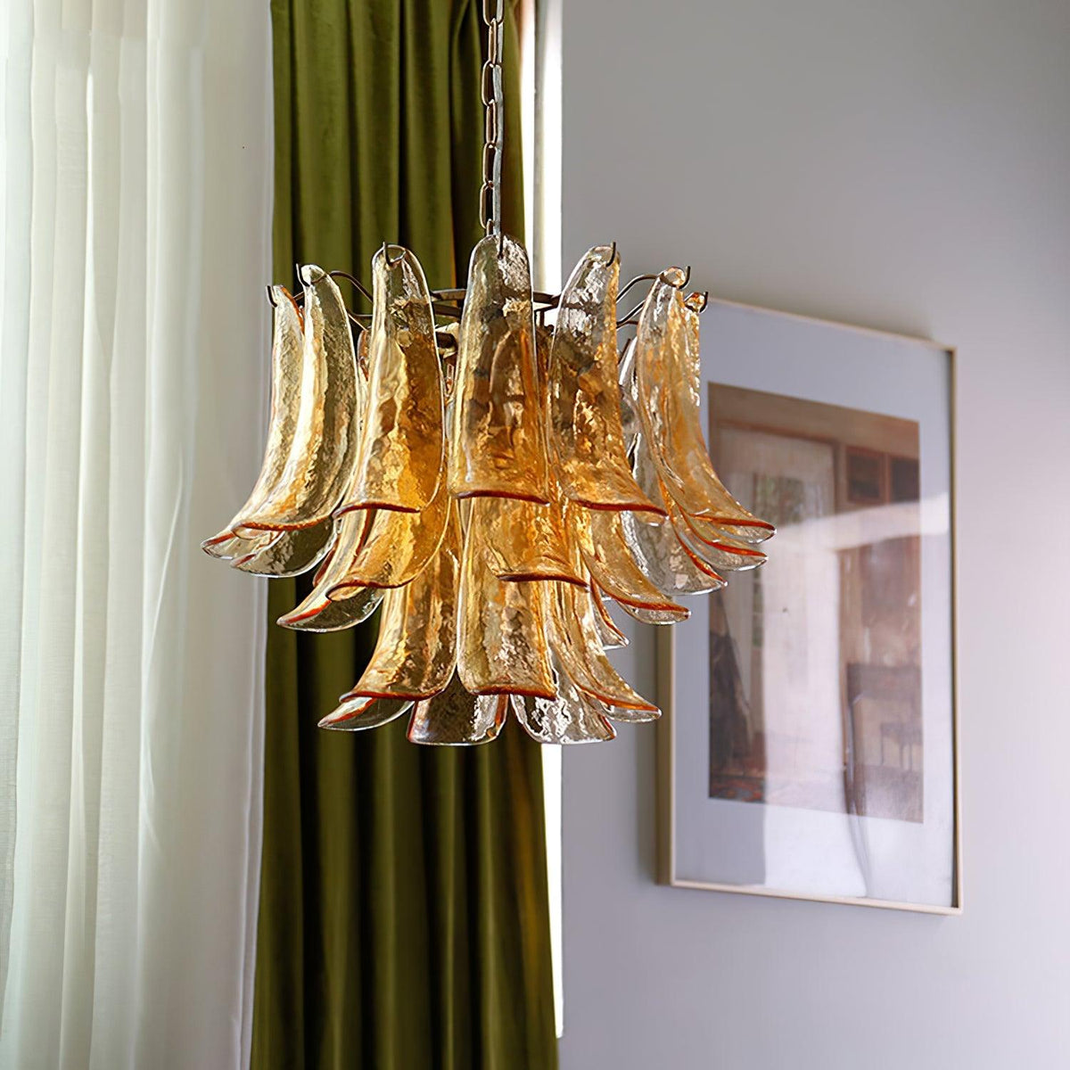 This Clérachic amber glass chandelier hangs in front of green and white curtains, with a framed print on the wall at its right.