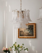 A pale glass leaf chandelier from Clérachic hangs high on a white wall, with a gold-framed townscape and pink roses below.