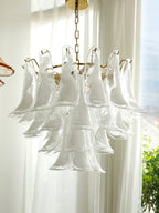Clear glass leaves overlap in tiers on this Clérachic chandelier, seen against bright sheer curtains and a window with greenery.