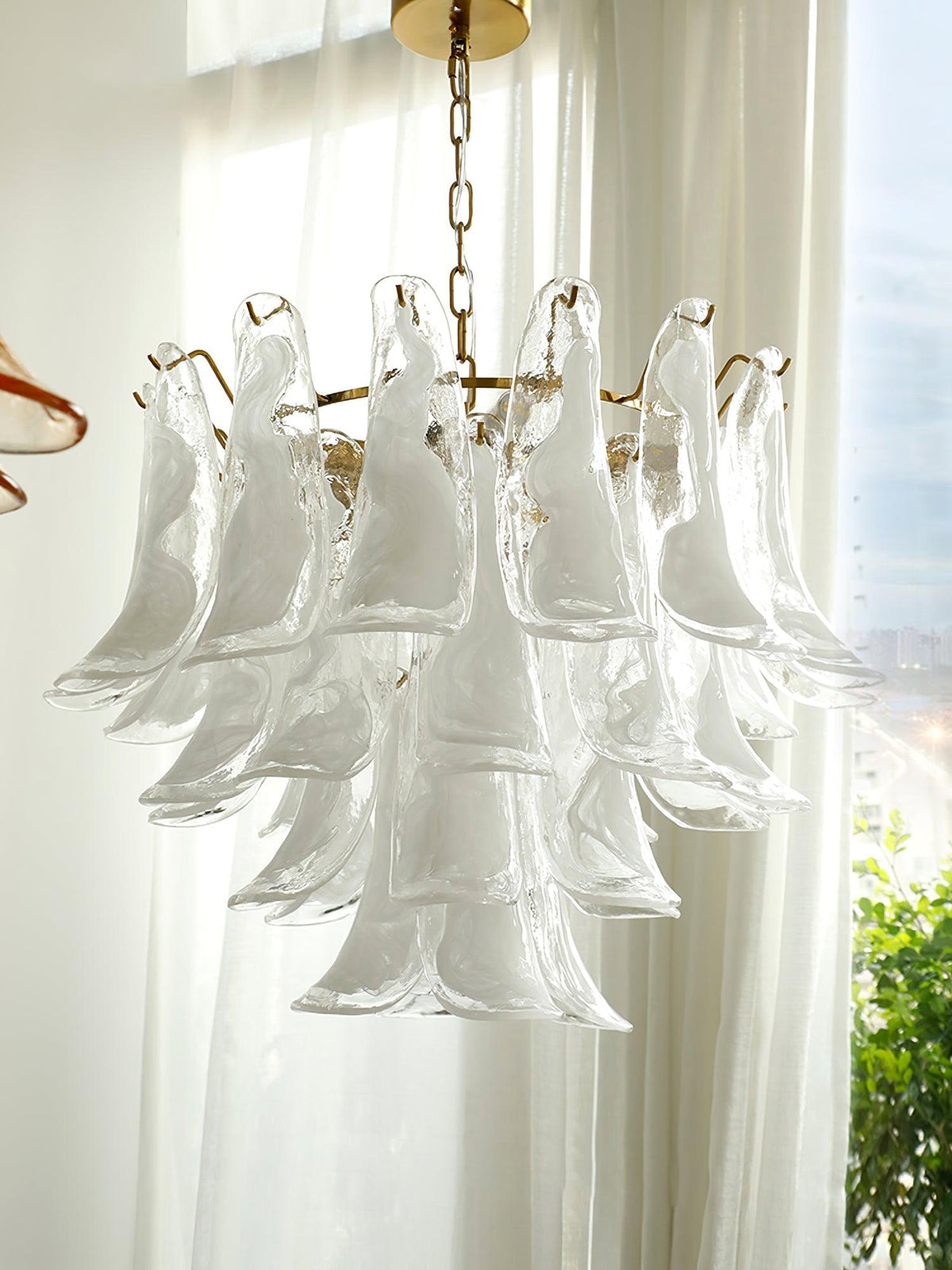 Clear glass leaves overlap in tiers on this Clérachic chandelier, seen against bright sheer curtains and a window with greenery.
