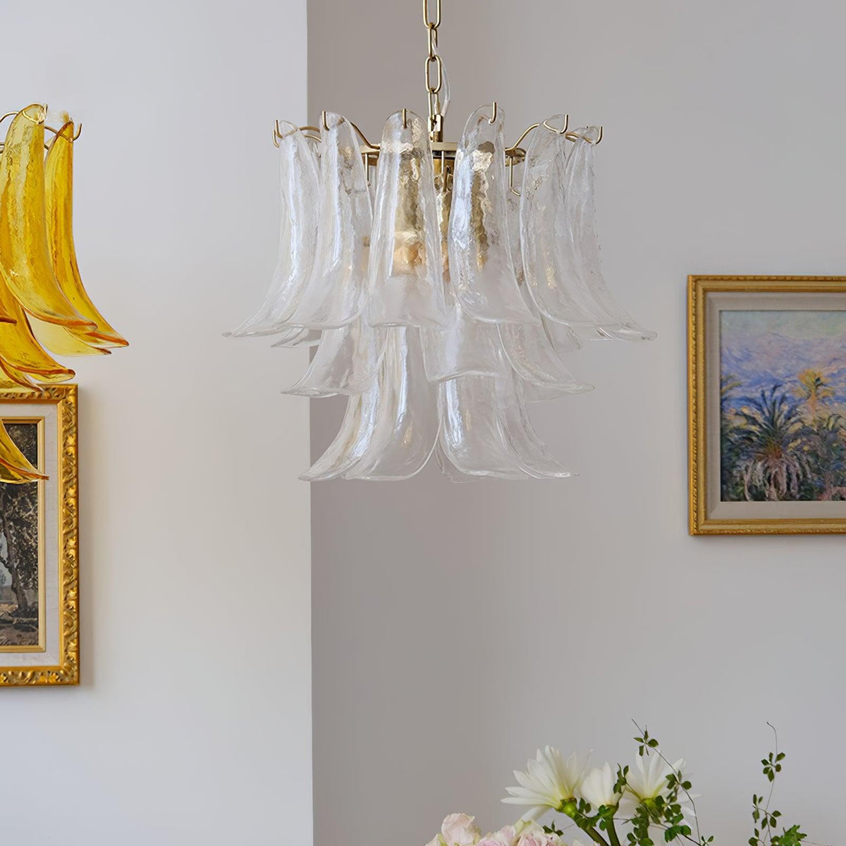 Clear glass leaf chandelier from Clérachic beside a gold-framed painting of palm trees, with white flowers at the bottom edge.