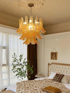 A lit amber glass leaf chandelier from Clérachic hangs in a bedroom with a tufted headboard, a knitted throw and sheer curtains.