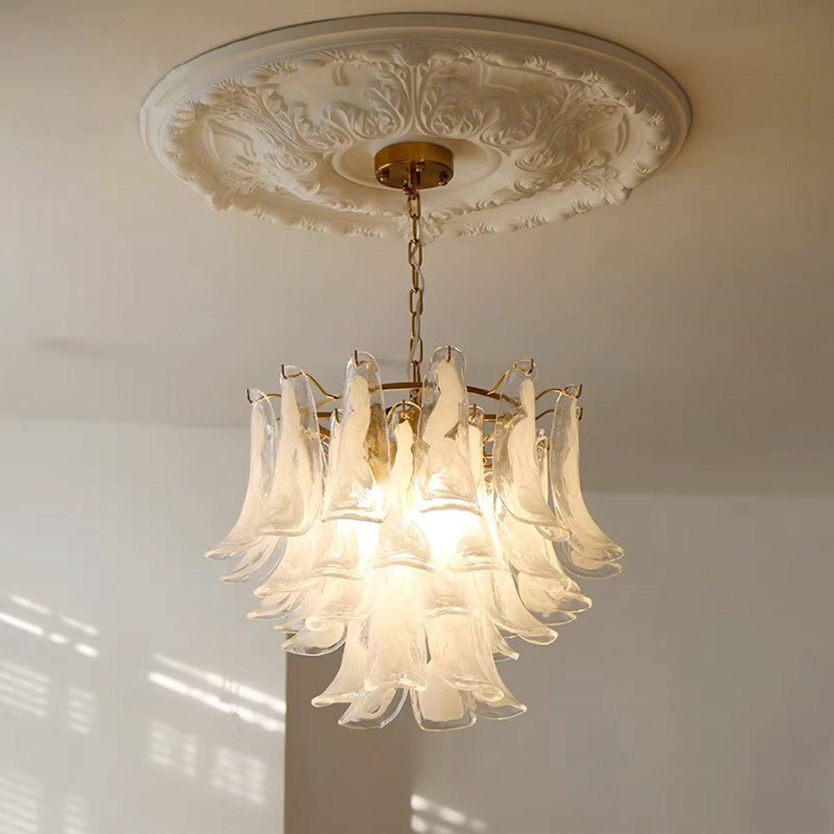 White cloud glass chandelier from Clérachic glowing under a plaster ceiling rose, with soft window light on the wall below.