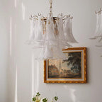 Frosted glass leaf chandelier from Clérachic hanging beside a gold-framed painting of a village street on a white wall.
