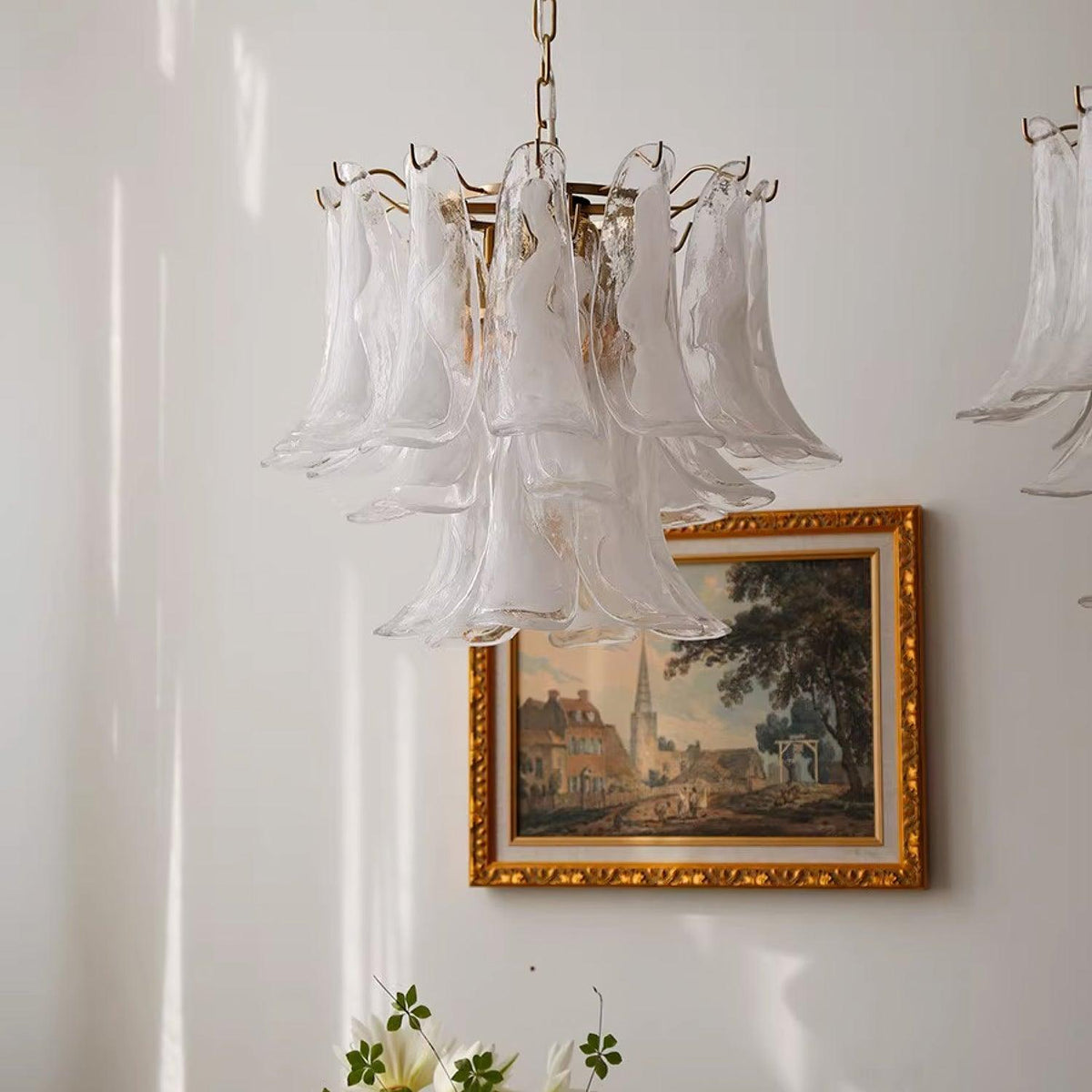 Frosted glass leaf chandelier from Clérachic hanging beside a gold-framed painting of a village street on a white wall.
