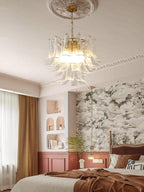 A lit clear glass leaf chandelier from Clérachic over a bedroom with a pink panelled wall, a brown throw and wallpaper.