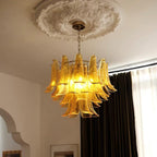 Warm amber glass leaves glow on this Clérachic chandelier, hanging under a plaster ceiling rose beside dark curtains and an arched doorway.