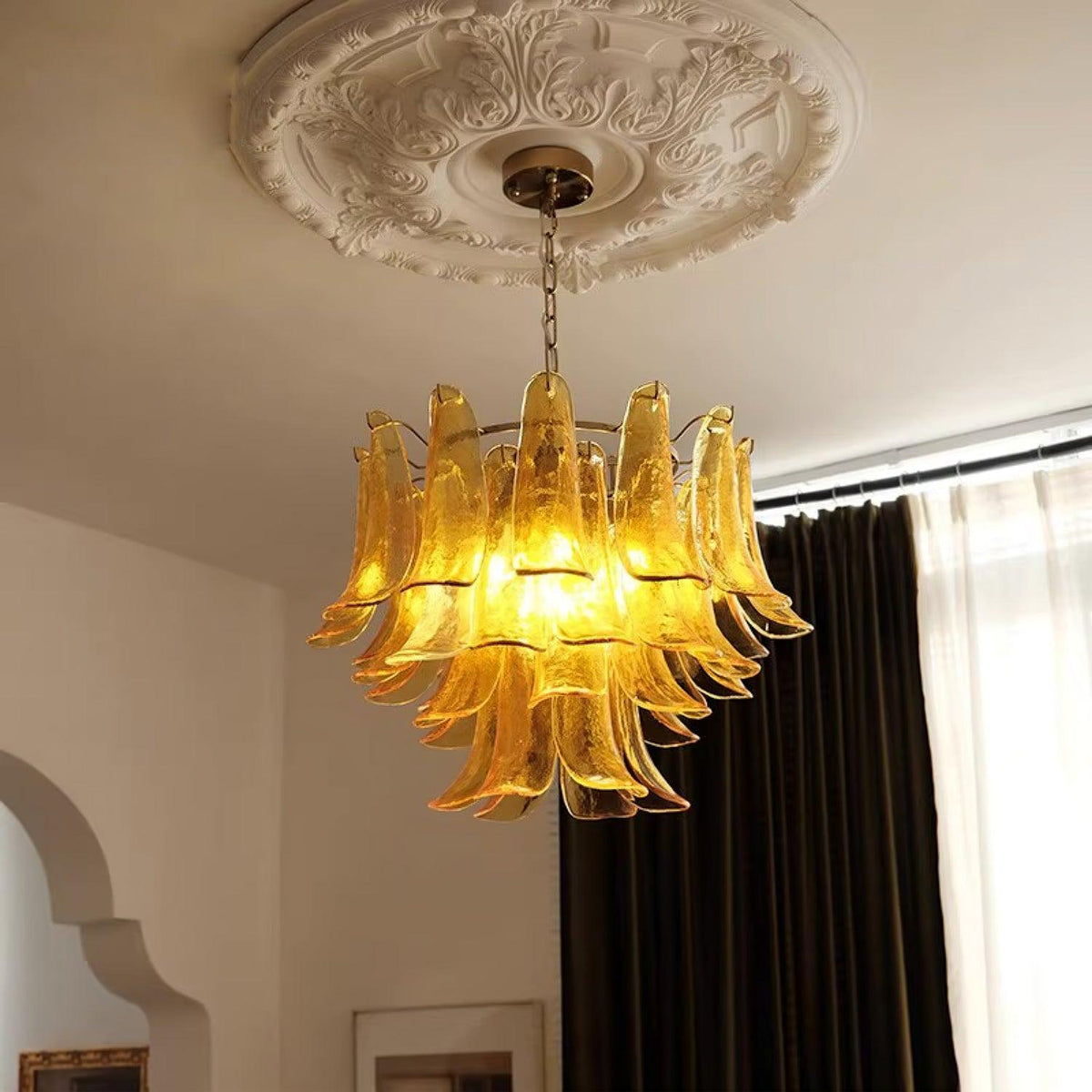 Warm amber glass leaves glow on this Clérachic chandelier, hanging under a plaster ceiling rose beside dark curtains and an arched doorway.