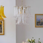 Pale clear glass leaf chandelier from Clérachic on a white wall, with a gold-framed palm painting and white blooms below it.