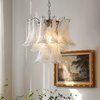 Frosted white glass leaves on a Clérachic chandelier, hanging near a gold-framed village painting with thin green stems below.