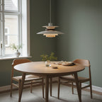 A pale Clérachic pendant hangs above a round oak table with wooden chairs, against a deep green wall.