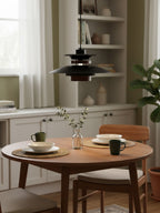 A black Clérachic pendant with a wood band hangs above a small round table set with bowls and mugs, near a bookshelf.