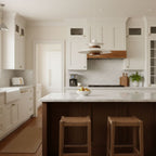 A pale Clérachic pendant hangs in a white kitchen, above a dark island with woven stools and a bowl of fruit.