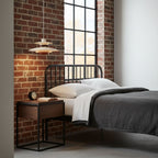 A cream Clérachic pendant with a wood band hangs by a brick wall, above a black-framed bed and a walnut side table.