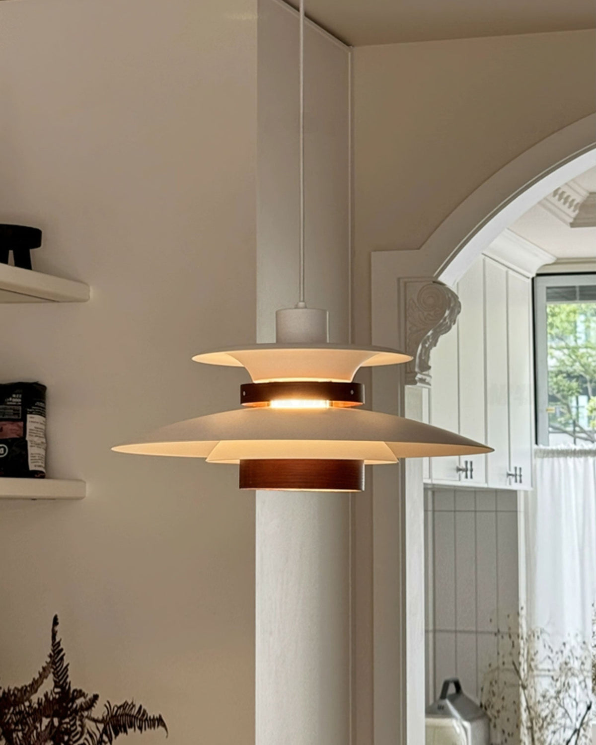 A grey-cream Clérachic pendant with a dark oak band hangs beside an arched doorway, with a dried plant at the bottom.