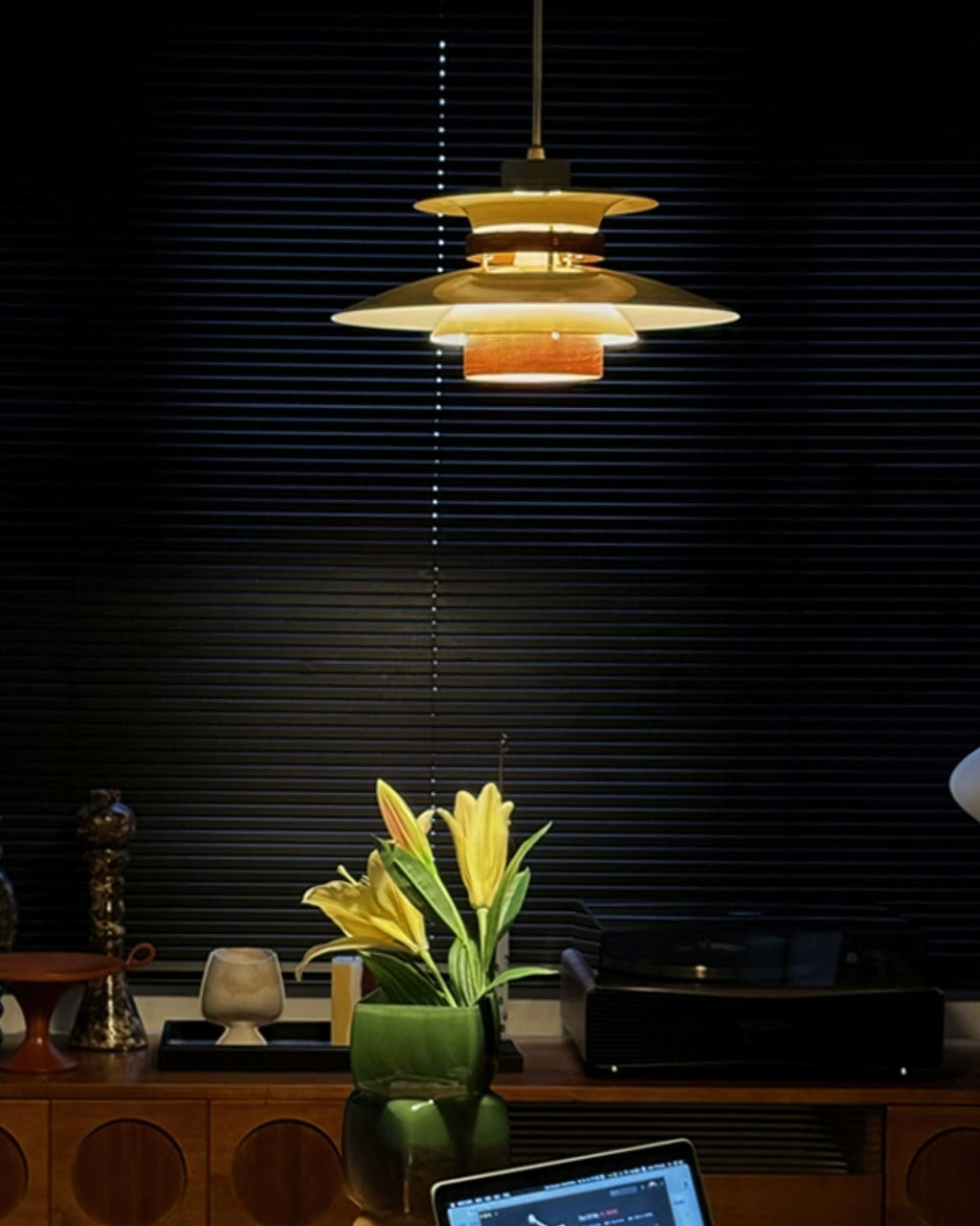 In a dark room with slatted blinds, a lit Clérachic pendant glows amber above a sideboard with yellow lilies.