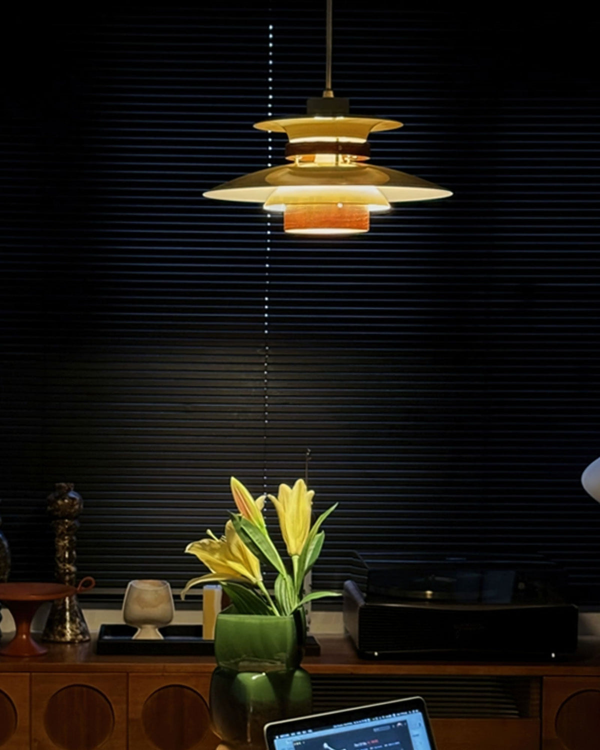In a dark room with slatted blinds, a lit Clérachic pendant glows amber above a sideboard with yellow lilies.
