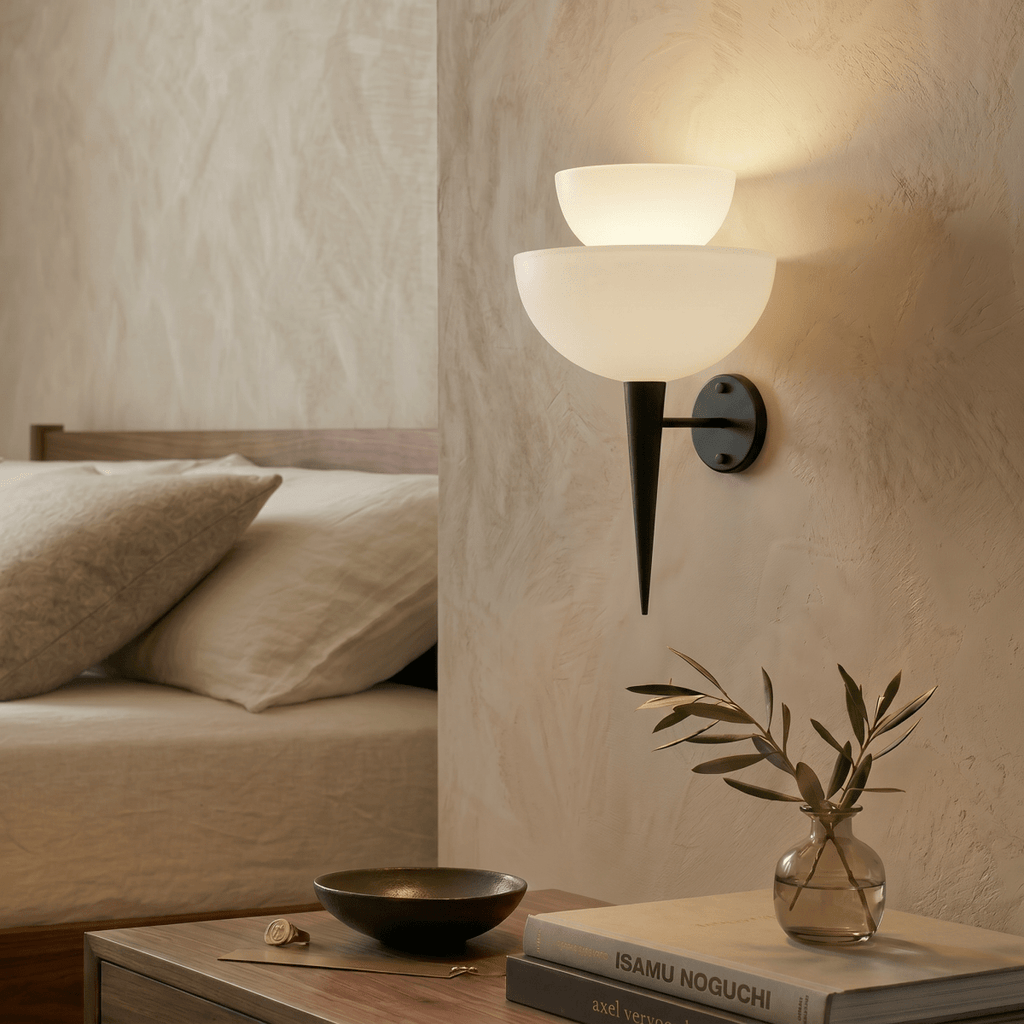 Clérachic wall lamp in matte black glowing over a nightstand with books and a small sprig of olive.