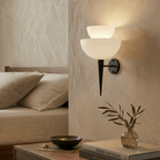 Clérachic wall lamp in matte black glowing over a nightstand with books and a small sprig of olive.