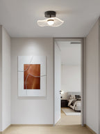 A single ceiling light with a wavy shade, glowing above a hallway with a framed copper print and an open bedroom door, from Clérachic.