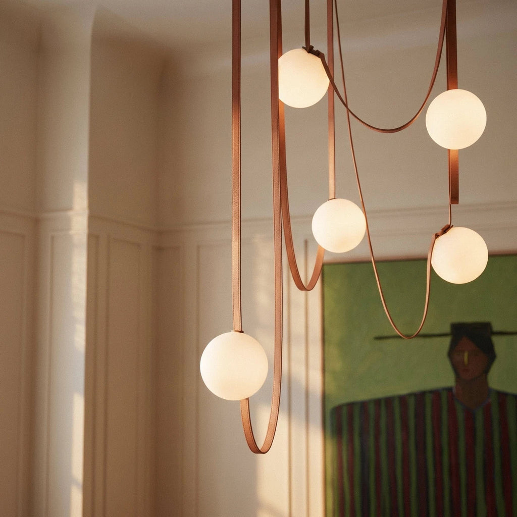 Close-up of a lit Clérachic chandelier's five frosted glass globes on looping leather straps above a dining table.