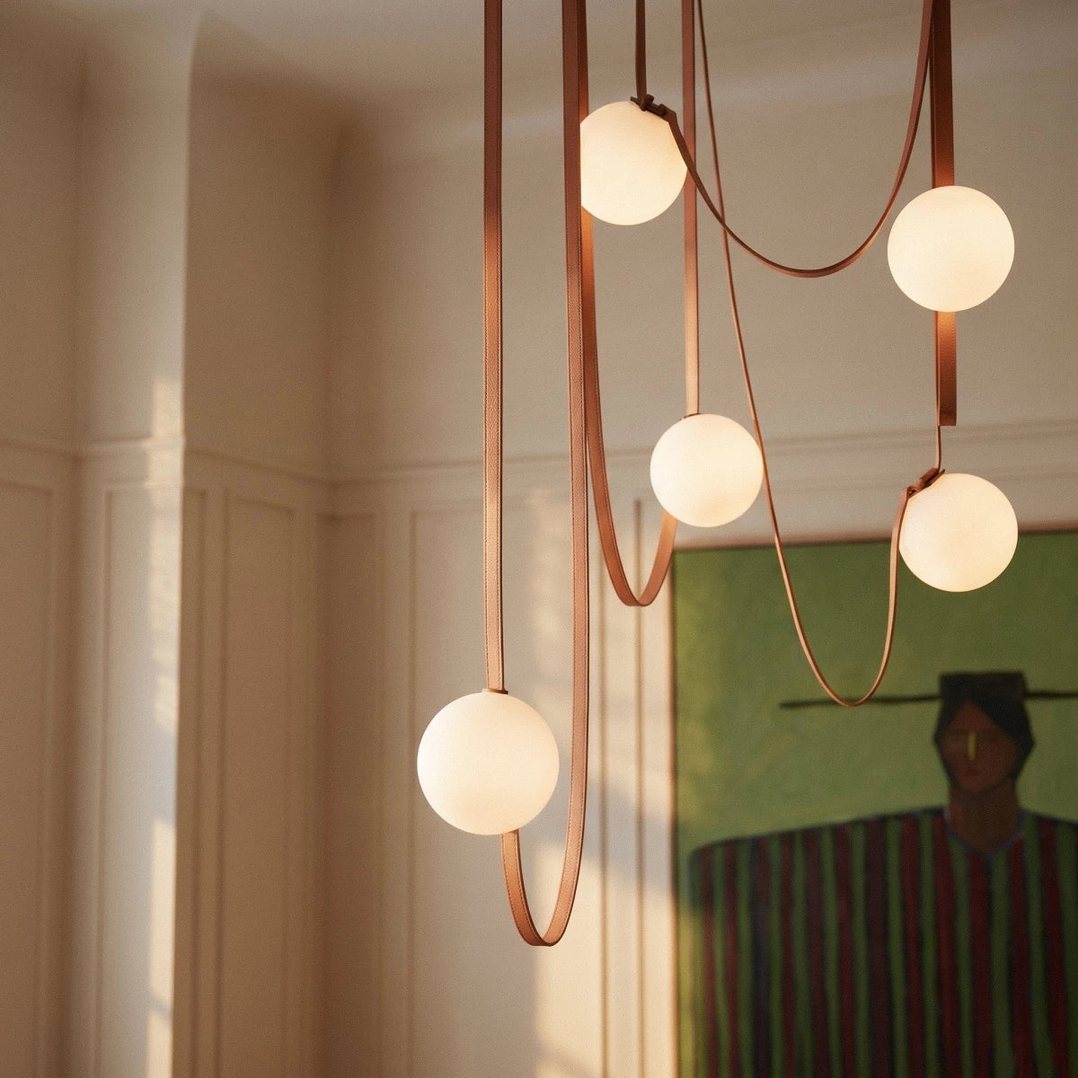 Close-up of a lit Clérachic chandelier's five frosted glass globes on looping leather straps above a dining table.
