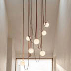 Close-up of a lit Clérachic chandelier's seven frosted glass globes trailing down leather straps in a sunlit hallway.