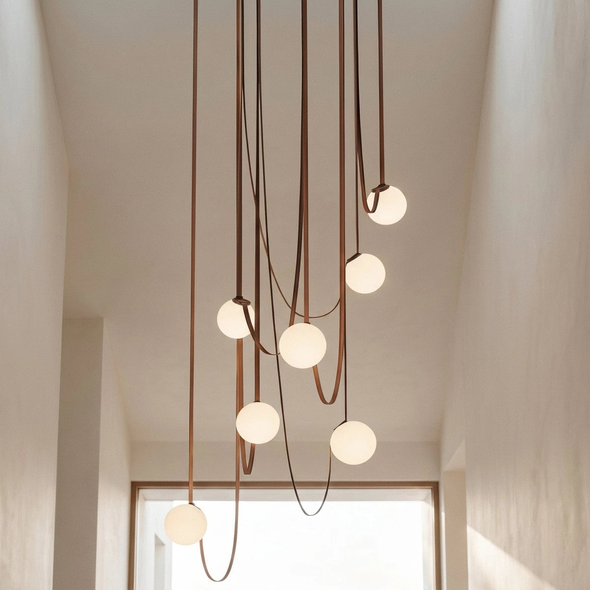 Close-up of a lit Clérachic chandelier's seven frosted glass globes trailing down leather straps in a sunlit hallway.