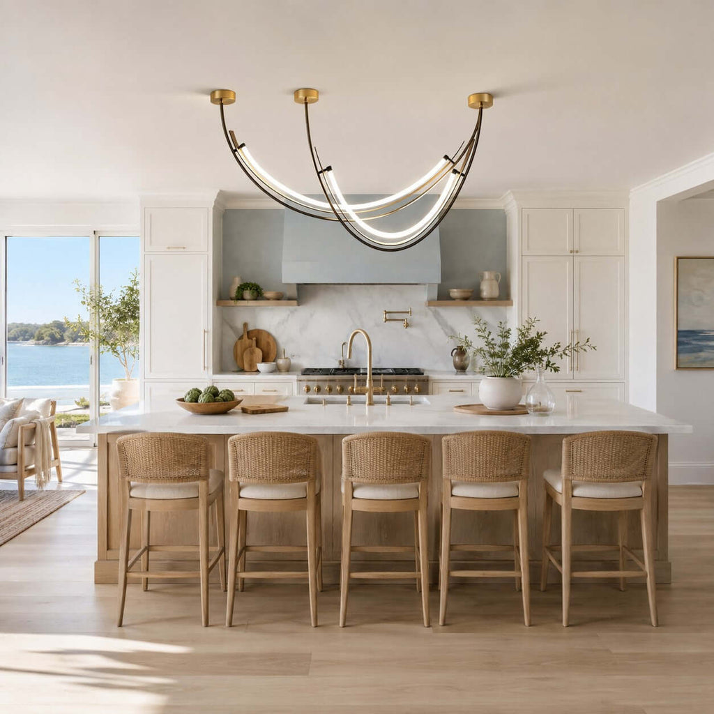 Lit arc linear pendant from Clérachic over a kitchen island with woven stools, with a sea view through the windows behind.
