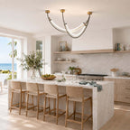 Arc pendant from Clérachic with glowing white strips, above a marble kitchen island and rattan stools, with a coastal view outside.