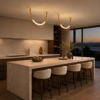 Two lit Clérachic loop pendants with gold caps hang above a marble kitchen island at dusk, with cream bar stools below.