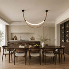 A black-capped Clérachic loop pendant, lit, hangs above a dark dining table in an open-plan room with a kitchen behind.